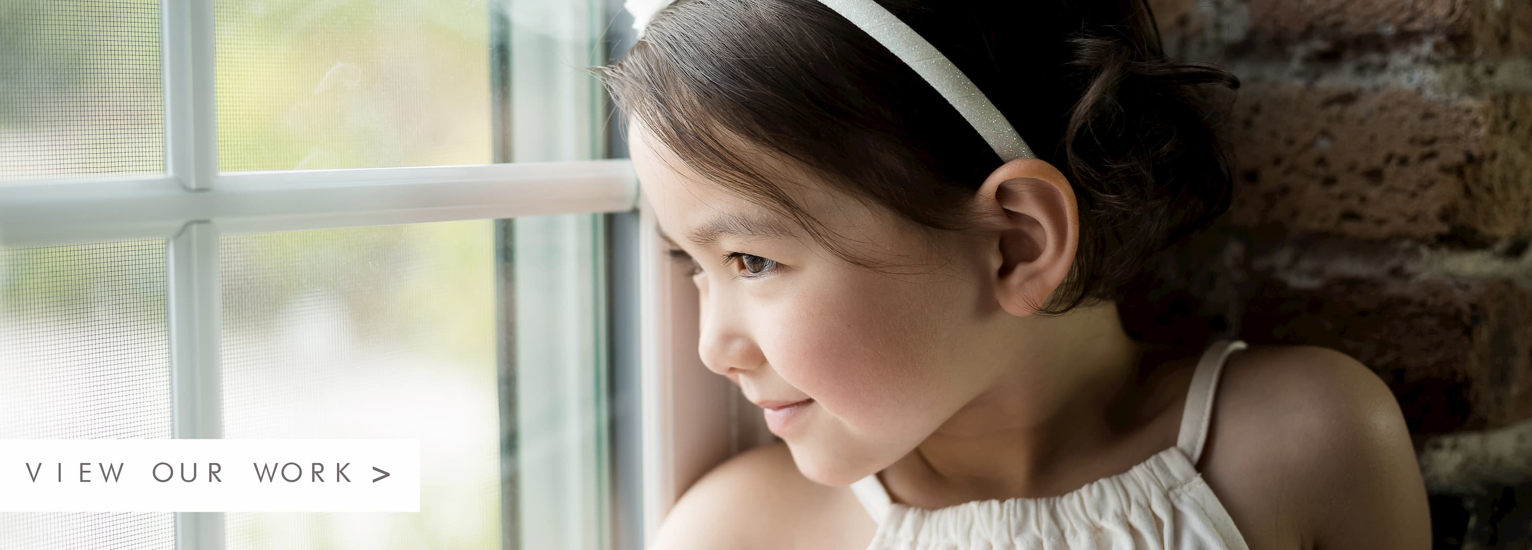 A child with rosy cheeks and a white headband in front of a brick wall looking out a window. To the left there is text to view our work
