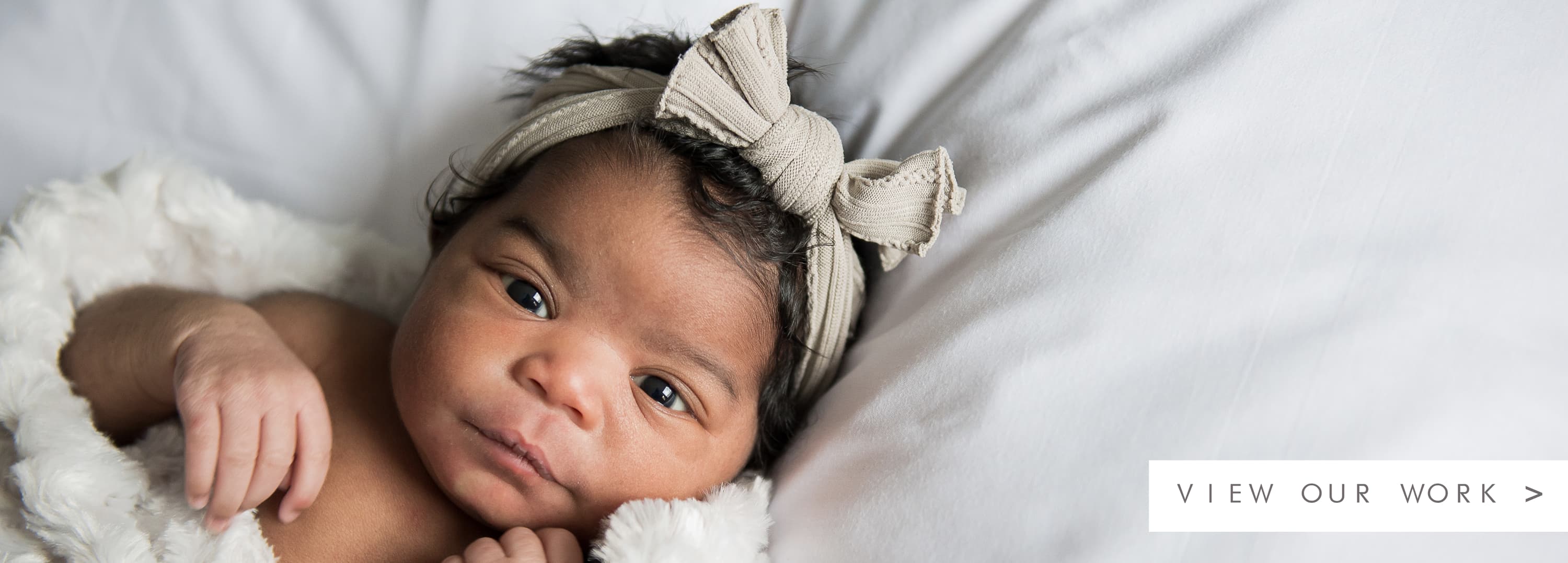 A blanket wrapped baby looking up with a brown bow on their head and text to view our work